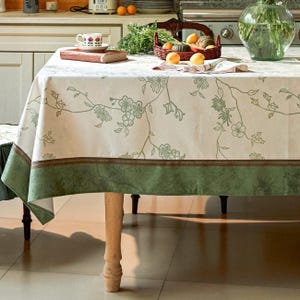 Könnte beinhalten: Eine rechteckige Tischdecke mit cremefarbener Basis, einem zarten grünen Blumenrankenmuster und einem einfarbigen dunkelgrünen Rand an der Unterkante. Der Stoff liegt auf einem Holztisch und zeigt ein klares und schlichtes Design.