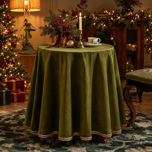 May include: A round table covered with a solid olive green velvet tablecloth featuring a decorative gold braided trim along the bottom edge. The table is set with a white ceramic teacup, a tall brass candle holder, and a festive floral arrangement.