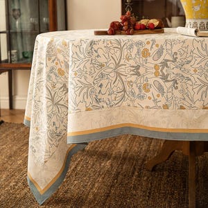 May include: A round tablecloth with a detailed floral pattern in cream, gray, and yellow. The border features a blue and gold trim. The tablecloth is draped over a wooden table, creating an elegant dining setting.