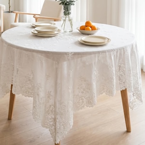 Puede incluir: Un mantel redondo blanco con intrincados bordados de encaje floral y un borde festoneado. La tela cubre una mesa circular, dispuesta con platos de cerámica color crema apilados y un cuenco que contiene tres naranjas frescas.