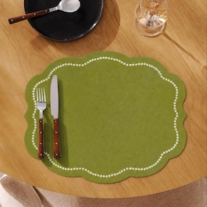 May include: An olive green scalloped placemat featuring a white dotted border. A stainless steel fork and a knife with brown handles are placed on the left side of the placemat. The placemat has a soft, textured surface and a rounded edge design.