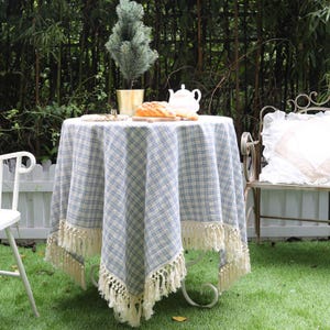 May include: A round table covered with a light blue and white plaid tablecloth featuring a decorative cream-colored fringe along the bottom edge. The table is set with a small potted evergreen tree, a white teapot, and a loaf of bread.