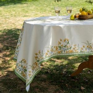 May include: A round white tablecloth featuring a decorative border of yellow flowers, green foliage, and small blue birds. The fabric is draped over a table, which holds two clear glass goblets and a wooden bowl filled with yellow lemons and limes.