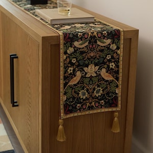 Op de afbeelding: Een decoratieve tafelloper met een zwarte achtergrond, versierd met vogels, bloemen en bladeren in groen, rood en beige. De loper heeft gouden kwastjes en ligt op een houten kast.