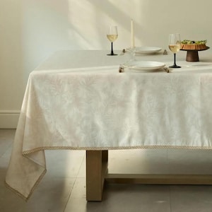 May include: A white tablecloth with a subtle leaf pattern covers a wooden table. Two wine glasses, plates, and a cake stand are set on the table. The tablecloth has a gold trim.