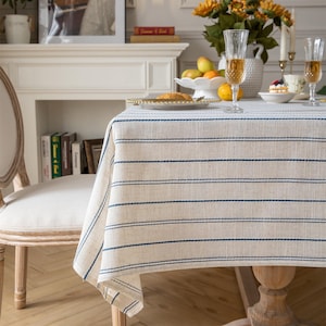 May include: A rectangular tablecloth featuring a woven beige fabric base with thin, horizontal navy blue stripes. The fabric has a visible textured weave and drapes over the edges of a wooden dining table, covering the surface completely.