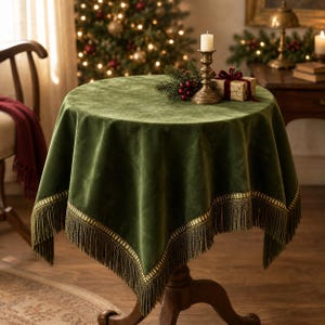 May include: A round, dark green velvet tablecloth featuring a gold-toned metallic trim and a long, matching fringe along the edges. The fabric has a soft, matte texture that drapes over the sides of a circular wooden pedestal table.
