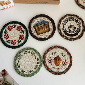 May include: Five round, braided coasters featuring various embroidered designs: a red floral wreath, a basket of blueberries, a coffee cup surrounded by beans, a green leafy vine, and an acorn with the text "WELCOME" in a red and green border.