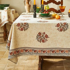 Könnte beinhalten: Beige Tischdecke mit floralem Muster in Braun- und Rottönen. Die Tischdecke hat einen dekorativen Rand mit sich wiederholendem Blumendesign und einem gestreiften Rand. Der Tisch ist mit Geschirr, Kerzen und Kürbissen gedeckt.