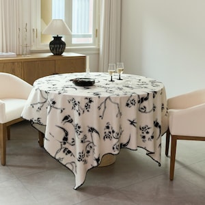 Puede incluir: Un mantel redondo con una base de color crema y un patrón repetitivo de flores y ramas negras. La tela cubre una mesa circular, la cual está dispuesta con dos copas de cristal transparente y una pequeña pila de platos negros en el centro.