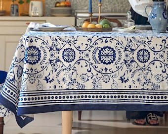 Puede incluir: Un mantel rectangular con base blanca y un patrón repetitivo de flores y volutas en azul oscuro. El diseño incluye un borde ancho con un motivo de líneas punteadas y un borde sólido en azul oscuro sobre una superficie de mesa de madera.