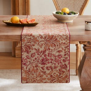 Puede incluir: Un camino de mesa rectangular con base color canela y un patrón floral de damasco repetitivo en rojo y naranja. El camino tiene un borde rojo sólido a lo largo de los lados largos y está colocado sobre una mesa de madera con un frutero.