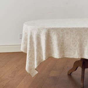 May include: An oval tablecloth in a light beige color featuring a subtle, repeating floral damask pattern woven into the fabric. The edges of the tablecloth are finished with a delicate, scalloped trim in a slightly darker shade of beige.