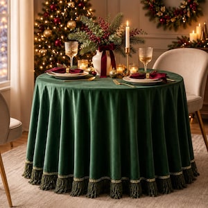 May include: A round dark green velvet tablecloth with a gold-trimmed fringe edge. The table is set with white plates, maroon napkins, gold napkin rings, ribbed glass tumblers, and a centrepiece of greenery, red berries, and candles.