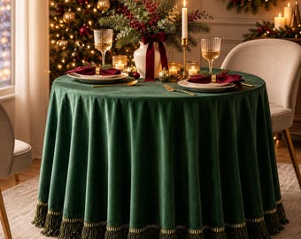 May include: A round dark green velvet tablecloth with a gold-trimmed fringe edge. The table is set with white plates, maroon napkins, gold napkin rings, ribbed glass tumblers, and a centerpiece of greenery, red berries, and candles.
