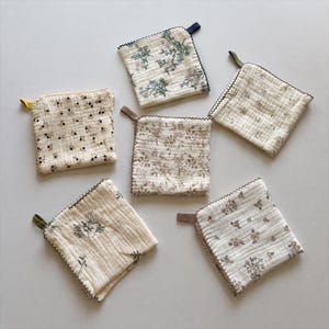 May include: Six square muslin cloths featuring various floral patterns in muted tones of blue, brown, and green. Each cloth has a textured, crinkled fabric surface, a scalloped edge finish, and a small fabric loop attached to one corner for hanging.