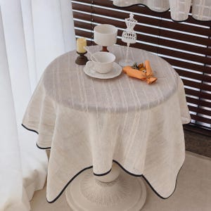 May include: A round table covered with a beige, vertically striped fabric featuring a dark trim along the scalloped edge. On the table sits a white ceramic cup and saucer, a small white candle, a white wire dress form, and a small orange bouquet.