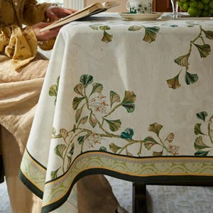 Op de afbeelding: Een crèmekleurig tafelkleed met een botanisch ontwerp van groene en bruine bladeren en witte bloemen. De rand heeft een groen en geel patroon. Een persoon leest een boek aan tafel.