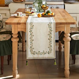 May include: A wooden dining table with a cream-coloured runner featuring a floral design. The runner is adorned with green and brown leaves and has green tassels at each end. The table is set with a cup, a plate of fruit, and a bowl of citrus fruits.