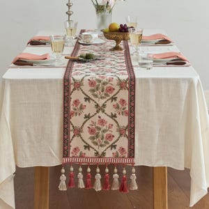 Puede incluir: Un camino de mesa rectangular con fondo crema y un patrón repetitivo de flores rosas y enredaderas verdes, enmarcado por un borde oscuro. El camino termina en una fila de borlas decorativas en tonos alternos de crema y rosa empolvado.
