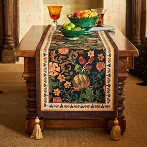 May include: A dark floral patterned table runner with a brown border and tassels. The runner features a black background with orange, yellow, and green flowers. The runner is placed on a wooden table with fruit-filled bowls and glasses.
