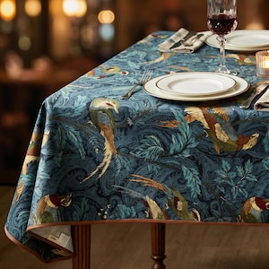 May include: A dark blue tablecloth featuring a repeating pattern of colorful pheasants and intricate swirling foliage. The fabric is draped over a table set with white ceramic plates, a silver fork, a wine glass, and a small candle holder.