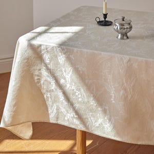 May include: A cream-coloured tablecloth featuring a subtle, tone-on-tone jacquard floral pattern. The fabric has a soft sheen and a finished hem. On the surface sits a small, metallic silver lidded bowl and a black metal candle holder.