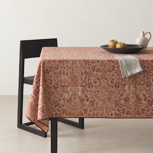 May include: A rectangular tablecloth featuring a repeating red and orange floral damask pattern on a light beige background. The fabric has a thin red border along the edge. A black chair, a dark bowl with two apples, and a white napkin are visible.