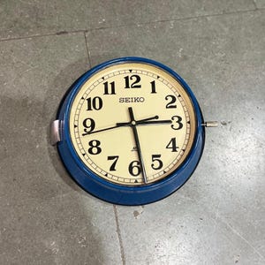 May include: A round Seiko wall clock with a blue frame and a cream-coloured face. Black numerals mark the hours, and black hands indicate the time. The clock has a silver winding knob and a silver metal bracket.