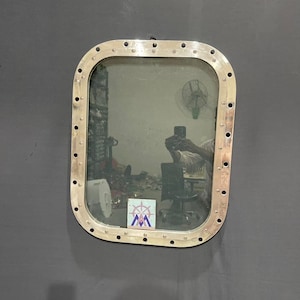 May include: A rectangular, porthole-style mirror with a silver metal frame and visible rivets. The mirror reflects a cluttered room with a fan, a chair, and a logo. The frame is mounted on a dark gray wall.