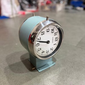 May include: A vintage, light blue Kappa clock with a silver-colored bezel. The clock face is white with black numerals and hands. The clock has a rectangular base and two small posts on top.