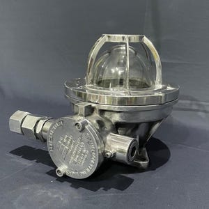 Pode incluir: Uma luminária industrial prateada com uma cúpula de vidro transparente protegida por uma gaiola de metal. A luminária tem um corpo cilíndrico com um conduto lateral e uma placa circular com texto estampado. Projetada para durabilidade e segurança.