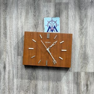 May include: A rectangular Seiko wall clock with a wood grain finish. The clock has silver-colored hour markers and hands. The clock face displays the Seiko brand name. A small, light blue and white nautical-themed image is above the clock.
