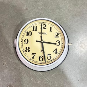 May include: A round Seiko wall clock with a cream-colored face and black numerals. The clock has a white metal frame and black hands. The word "SEIKO" is printed at the top of the clock face. The clock is on a gray surface.
