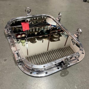 May include: A rectangular, polished silver mirror with a porthole design. The mirror has a thick, rounded frame with visible rivets and four circular handles. The reflection shows a cluttered shelf with various items.