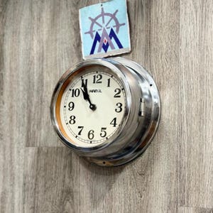 Op de afbeelding: Een ronde, zilverkleurige klok met een witte wijzerplaat en zwarte cijfers. De klok heeft een scharnierende rand en is aan een houten muur bevestigd. De wijzerplaat leest "HAN IL". Boven de klok staat een klein kunstwerk met een nautisch thema.