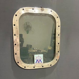 May include: A rectangular, silver-colored porthole window with rounded corners, mounted on a gray wall. The window's frame is secured with numerous rivets. The glass reflects a room with a fan and a sticker with a nautical emblem.