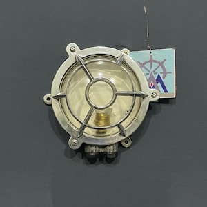 Op de afbeelding: Een ronde, zilverkleurige nautische lamp met een heldere glazen lens en een beschermende metalen kooi. De lamp heeft een messingkleurige lamp erin en is gemonteerd op een donkergrijze ondergrond. Een klein label met een nautisch logo is bevestigd.