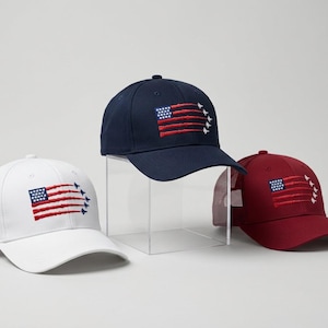 May include: Three baseball caps in white, navy blue, and maroon. Each cap features an embroidered design of the American flag where the stripes are formed by jet contrails and the stars are replaced by a formation of small white jet silhouettes.