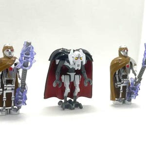May include: Three action figures from the Star Wars franchise. Two figures wear brown capes and helmets, holding purple weapons. The central figure is General Grievous, with a black cape and a white and gray body. The figures are on a white background.