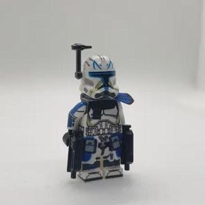Pode incluir: Uma minifigura Lego Star Wars Clone Trooper. A figura é predominantemente branca com detalhes azuis no capacete, armadura e acessórios. Possui uma viseira preta e uma arma preta. A figura está em pé.