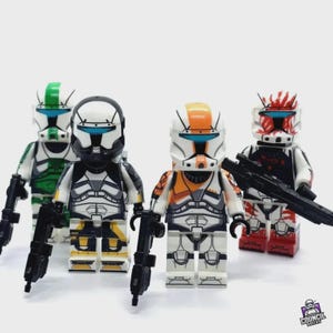 Peut inclure: Quatre figurines LEGO personnalisées de soldats clones, chacune avec des designs de casque et des motifs d'armure uniques. Les figurines sont principalement blanches, avec des accents de vert, de noir, d'orange et de rouge. Chaque figurine tient un fusil blaster noir.