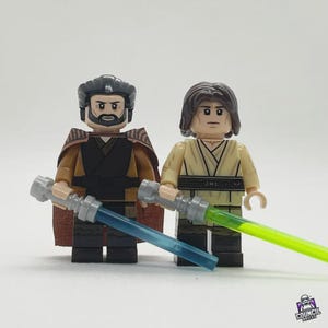 May include: Two Lego minifigures, one with a brown cape and a blue lightsaber, the other with a tan outfit and a green lightsaber. The figures stand side-by-side against a white background. The Council Bricks logo is in the bottom right corner.