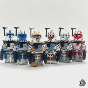 Può includere: Sei personaggi giocattolo di soldati clone di fantascienza, in vari colori, tra cui blu, arancione, rosso e grigio, con accessori neri. I personaggi sono in piedi.