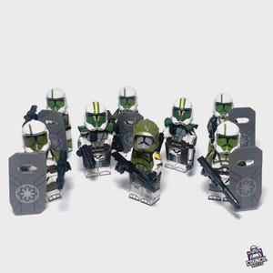 May include: A group of Lego minifigures, featuring white and green helmets and armor. Some figures hold black blasters and gray shields with a starburst design. The figures are from the Council Bricks brand.