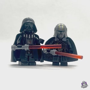 Könnte beinhalten: Zwei Lego-Figuren von Darth Vader und einer anderen Figur, beide in schwarzer Kleidung, halten rote Lichtschwerter. Die Darth-Vader-Figur hat einen schwarzen Helm und Umhang. Die andere Figur hat einen grauen Helm und Umhang. Der Hintergrund ist schlicht weiß.