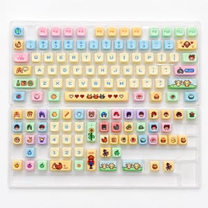 May include: A set of colorful keyboard keycaps with various icons and symbols. The keycaps feature pastel colors and include images of animals, food, and other decorative elements. The keyboard keys are arranged in a standard QWERTY layout.