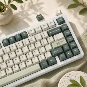May include: A white mechanical keyboard with green and off-white keycaps. The keyboard has a standard layout with function keys, arrow keys, and a number pad. The keycaps have a minimalist design with clear lettering. The keyboard is on a light-colored surface.