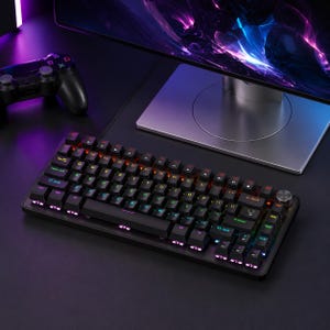 Puede incluir: Un teclado mecánico negro con teclas iluminadas. Las teclas están iluminadas con varios colores y una perilla plateada está en la esquina superior derecha. Un mando de juegos y un monitor de ordenador están en el fondo.