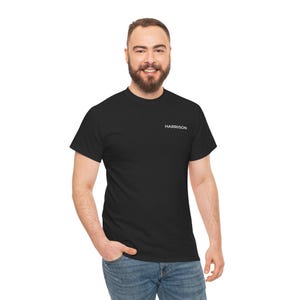 May include: Black t-shirt with the name "HARRISON" printed in white on the upper left chest. The shirt is a crew neck style and is being worn with blue jeans. The person is smiling.